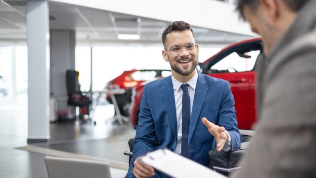 A smiling executive in a suit talks with a customer holding paperwork with cars in the background