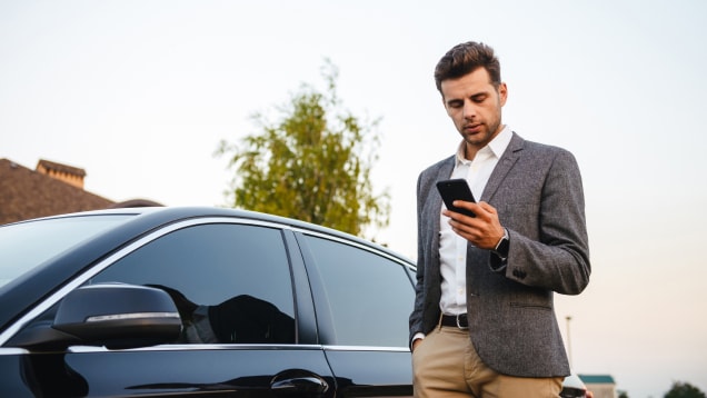Businessman wearing suit, standing near his luxury black car and using smartphone while holding in hand