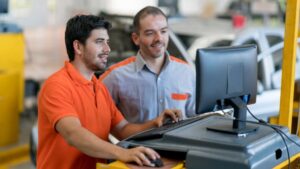 Two men collaborate on a computer inside a car repair shop