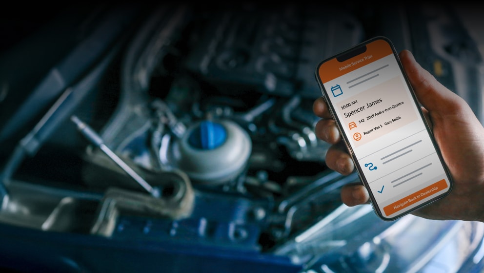 Hand holding a smartphone displaying the myKaarma platform in front of an open car hood, showing the engine compartment